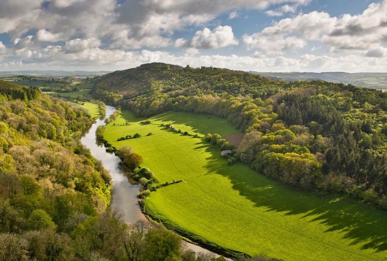 A winding river flows through a green valley with rolling, tree-covered hills under a cloudy sky.
