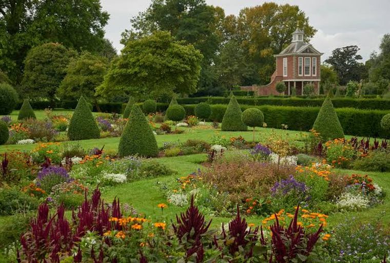 A formal garden with manicured trees, flower beds, and a small building in the background.