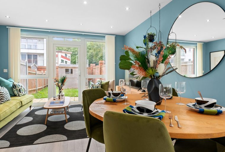 A dining and living room area with a green sofa, olive green chairs, a wooden dining table, and a large round mirror.