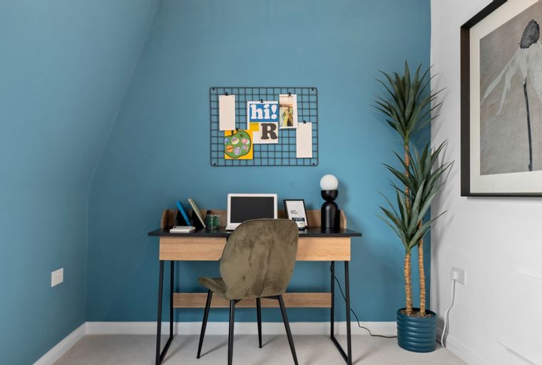 A desk with a laptop, chair, and a bulletin board on a blue wall.