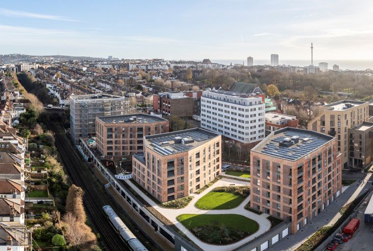 An aerial view of a modern apartment complex with brick buildings and green spaces, next to a railway line and overlooking a city and the sea.