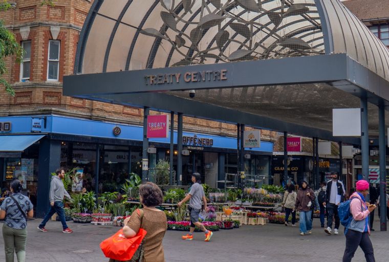A street scene with people walking past shops and a flower market under a covered walkway.