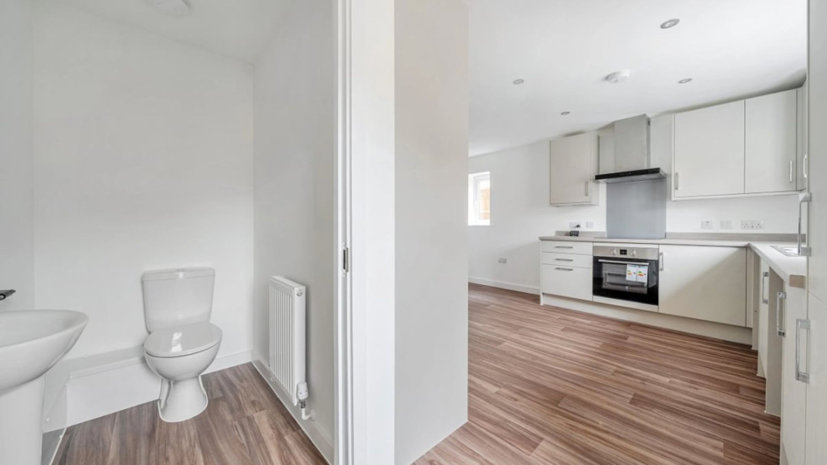 A toilet and sink in a room adjacent to a kitchen with white cabinets and an oven.