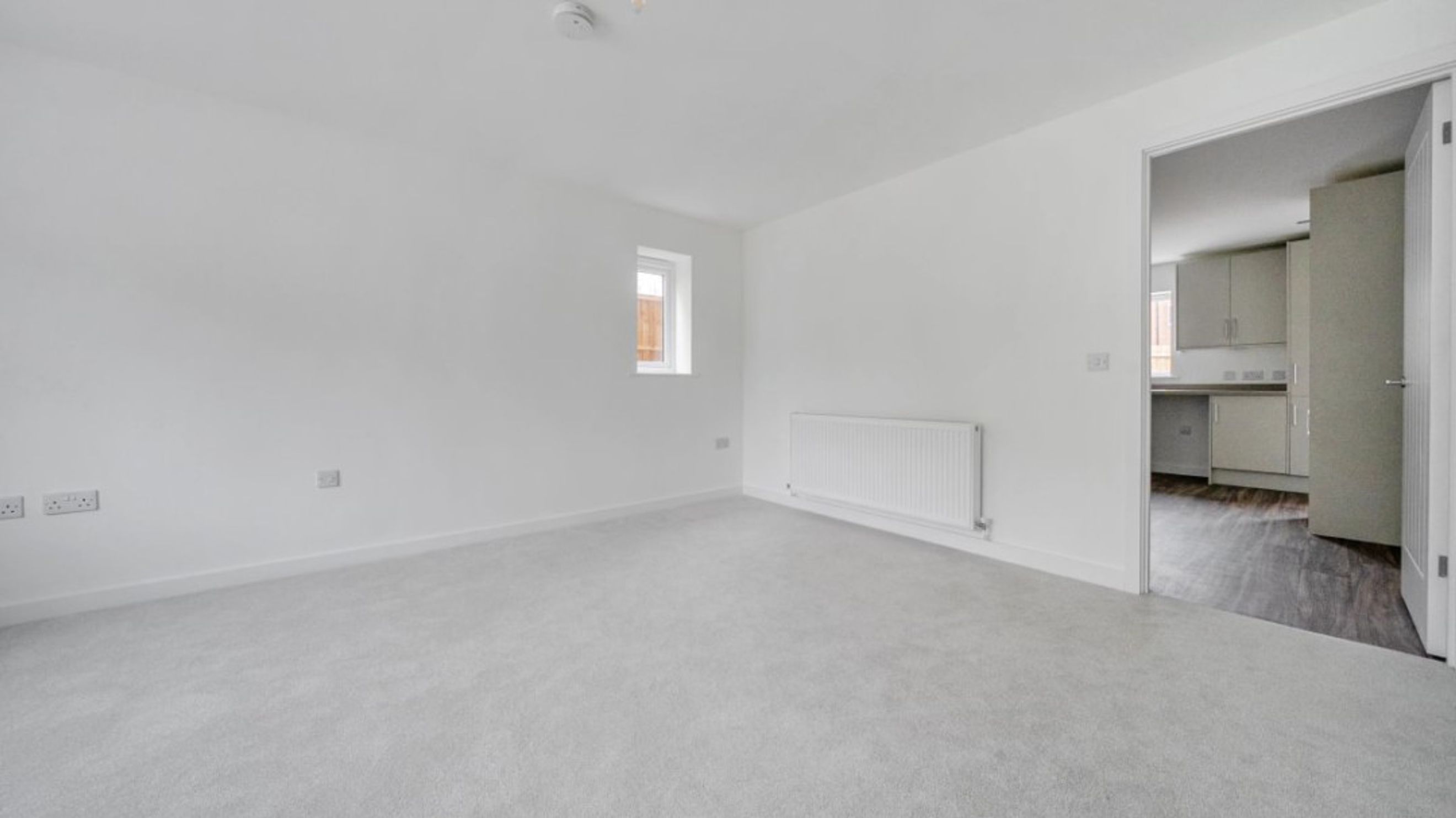 A living room with grey carpet, white walls, a window, and a radiator, with a view into a kitchen.
