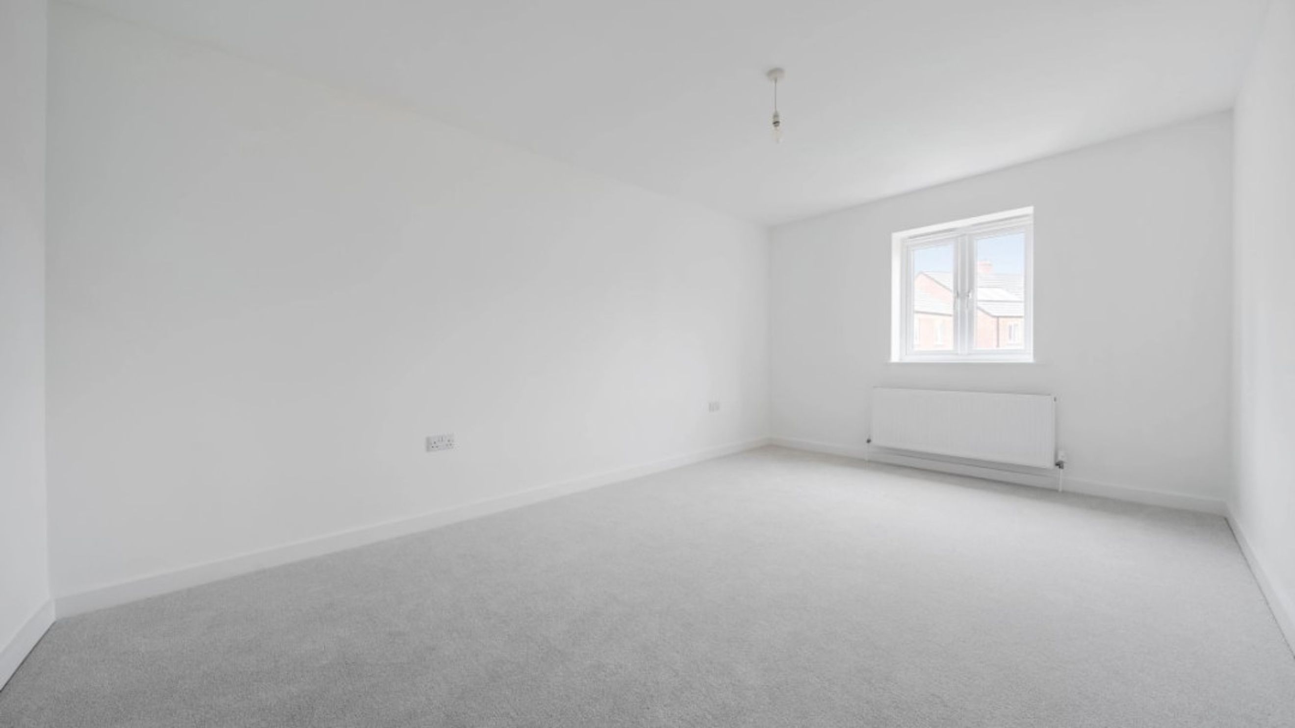 A spacious, empty room with grey carpet, white walls, and a window with a radiator.