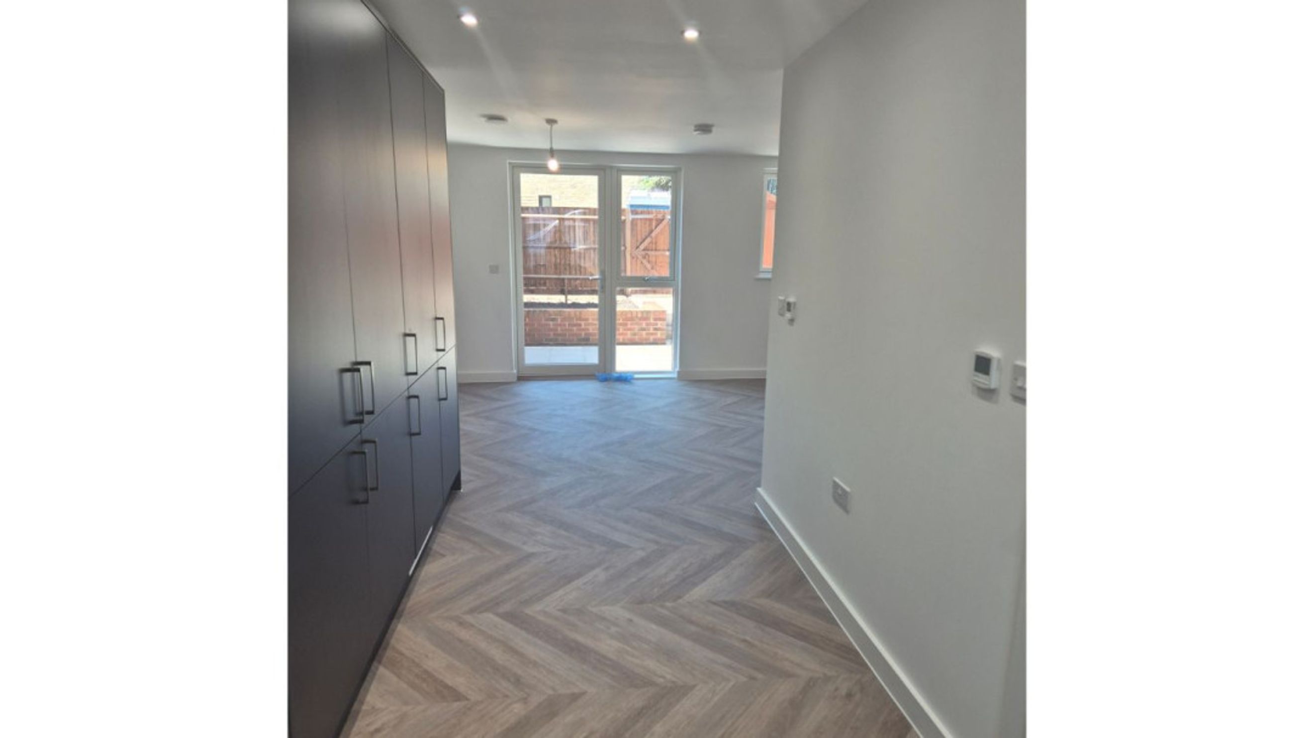 A hallway with a large dark grey wardrobe and a herringbone floor leading to a room with double glass doors.