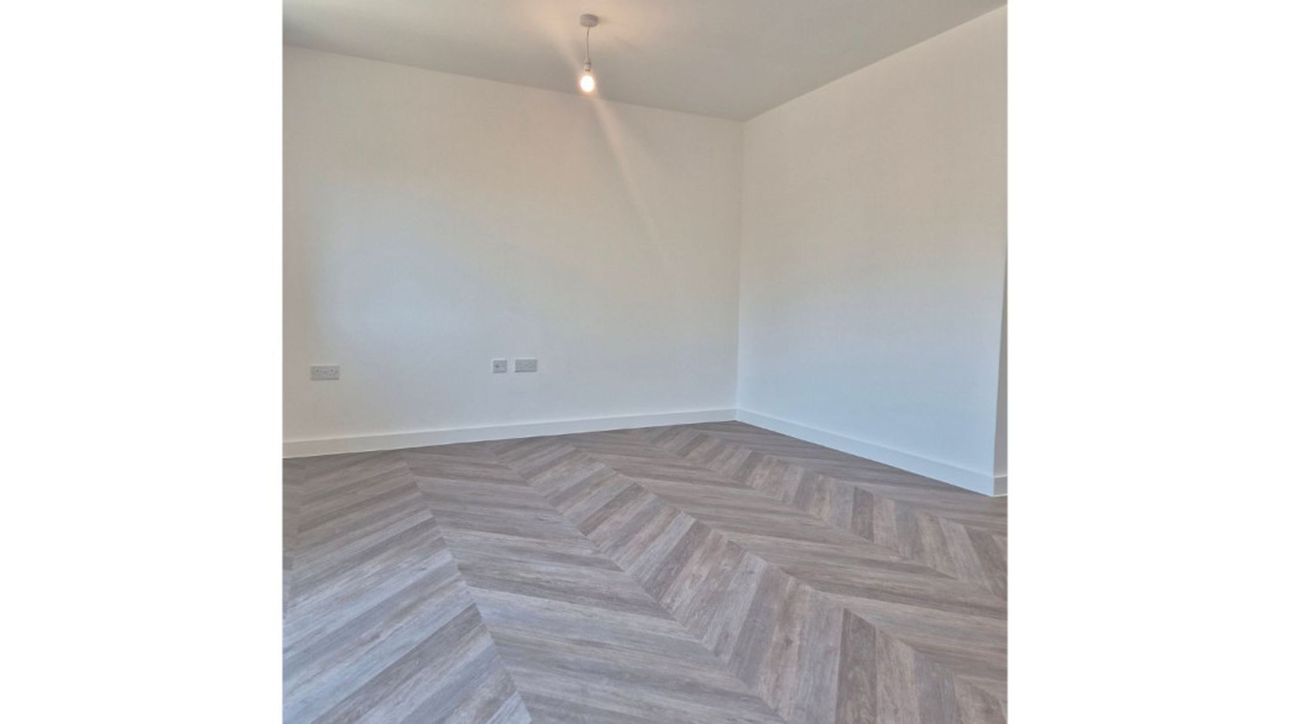 A corner of an unfurnished room with white walls and a herringbone wood-effect floor.