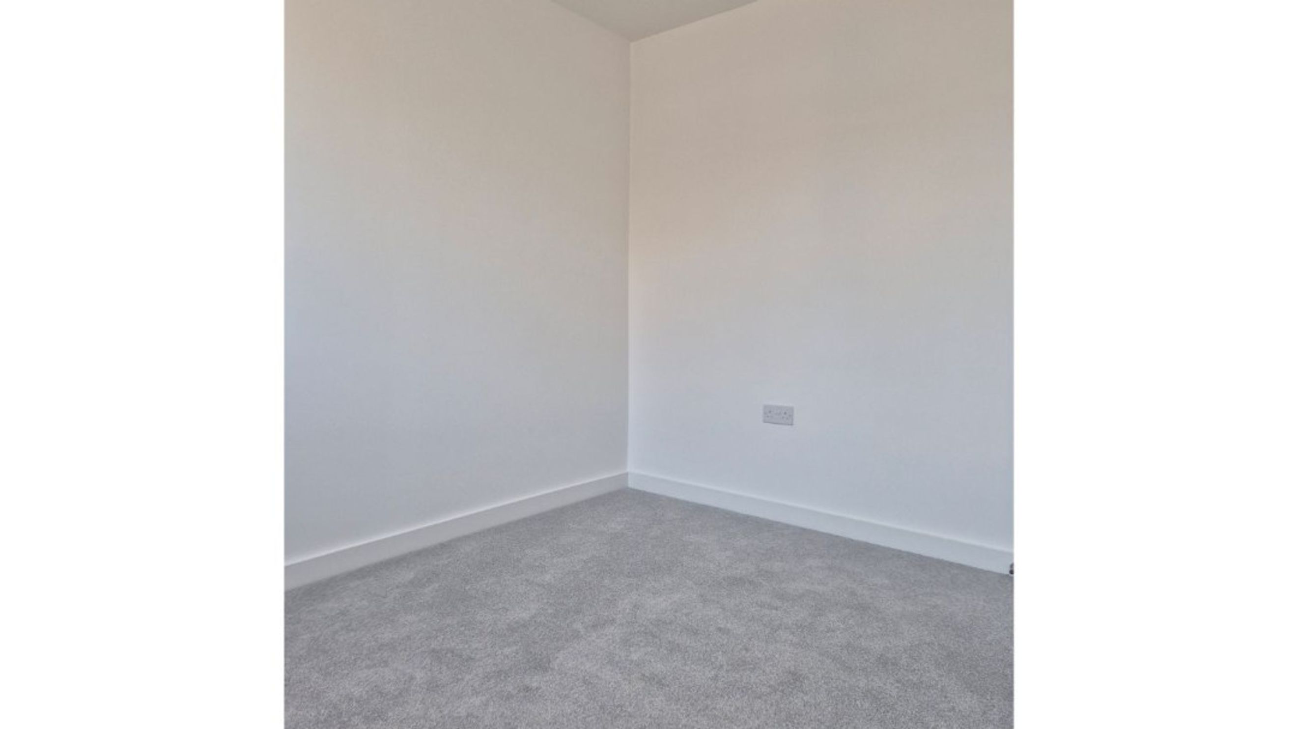 A corner of a room with white walls, grey carpet, and a white electrical socket.