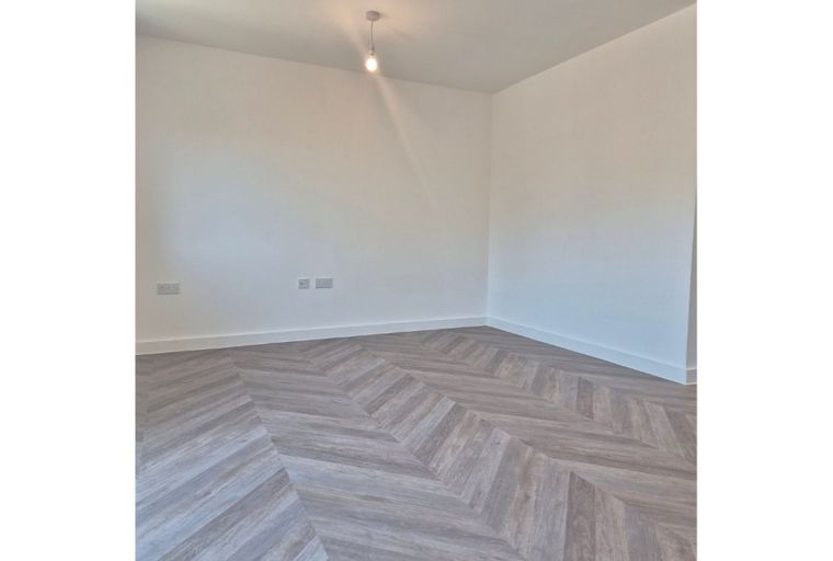 A corner of an unfurnished room with white walls and a herringbone wood-effect floor.