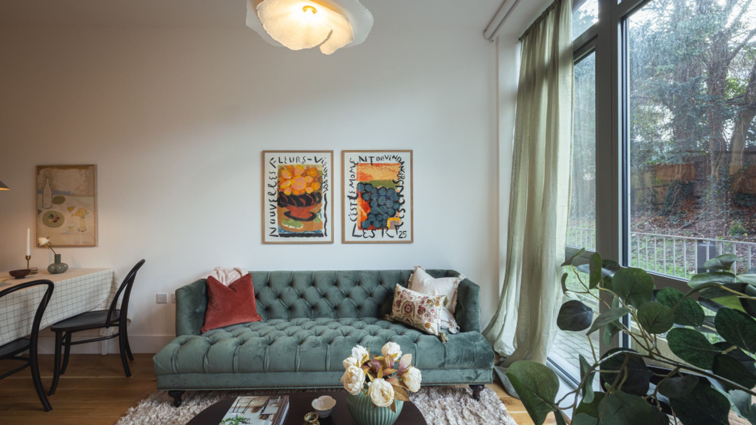 A living room with a green tufted sofa, two framed artworks, and a large window.