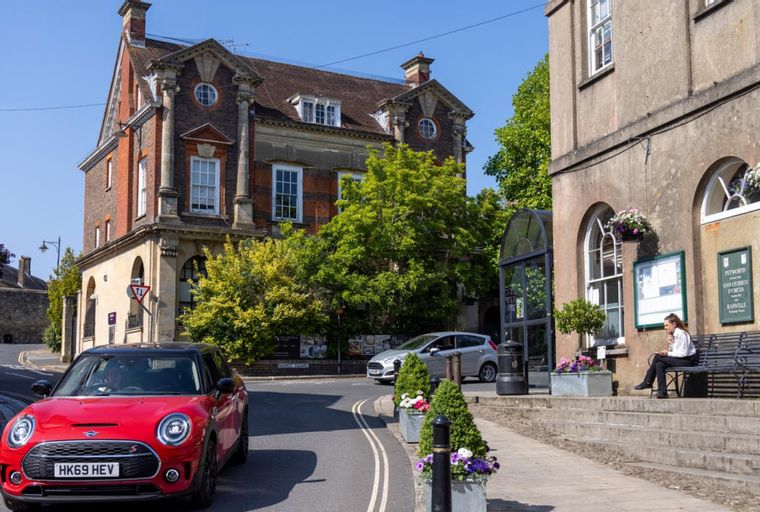 A red Mini Cooper drives down a street in front of historic buildings with a woman sitting on a bench.