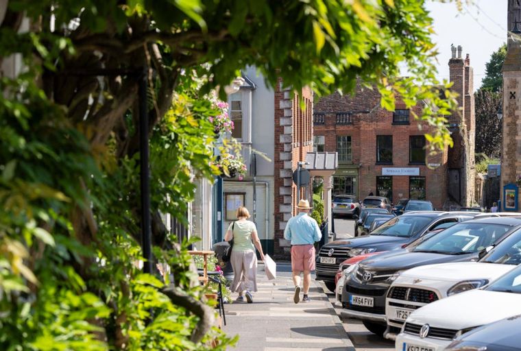 A street scene with people walking on a sidewalk next to parked cars and buildings.
