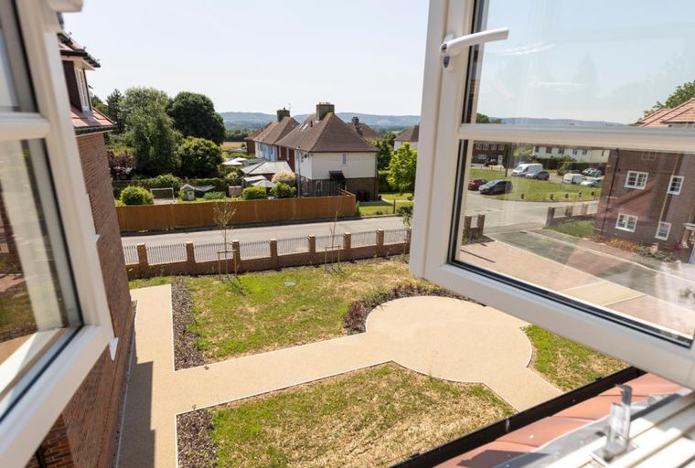 A view of a residential neighborhood through an open window, showing houses, gardens, and a street.