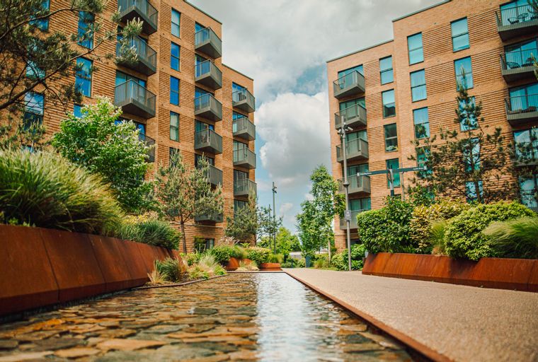 A view of modern brick apartment buildings with balconies, surrounded by greenery and a water feature.