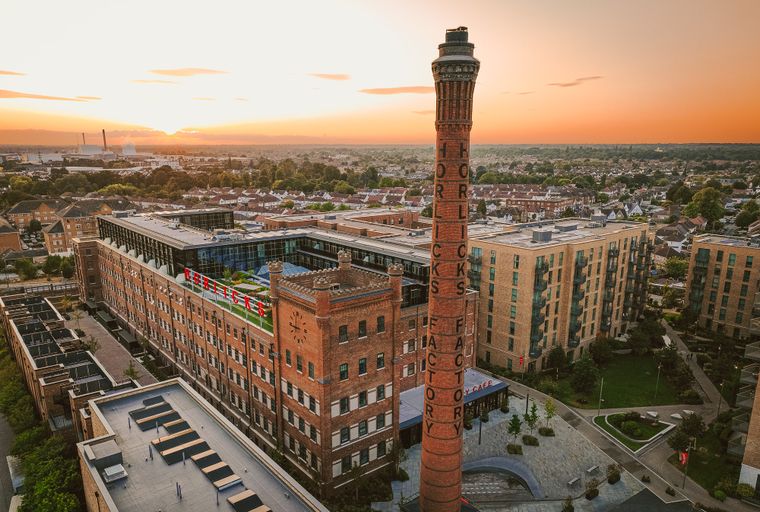 An aerial view of the Horlicks Factory complex at sunset, with a tall brick chimney and surrounding apartment buildings.