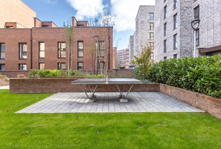 A ping pong table sits on a paved patio surrounded by a lawn and brick planters, with apartment buildings in the background.