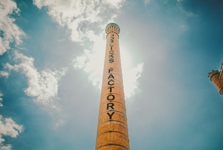 A tall brick chimney with the words 'HORLICKS FACTORY' written vertically on it, against a cloudy blue sky.