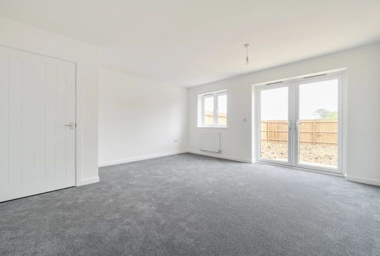 A spacious, unfurnished room with grey carpet, white walls, a white door, a window, and double doors leading outside.