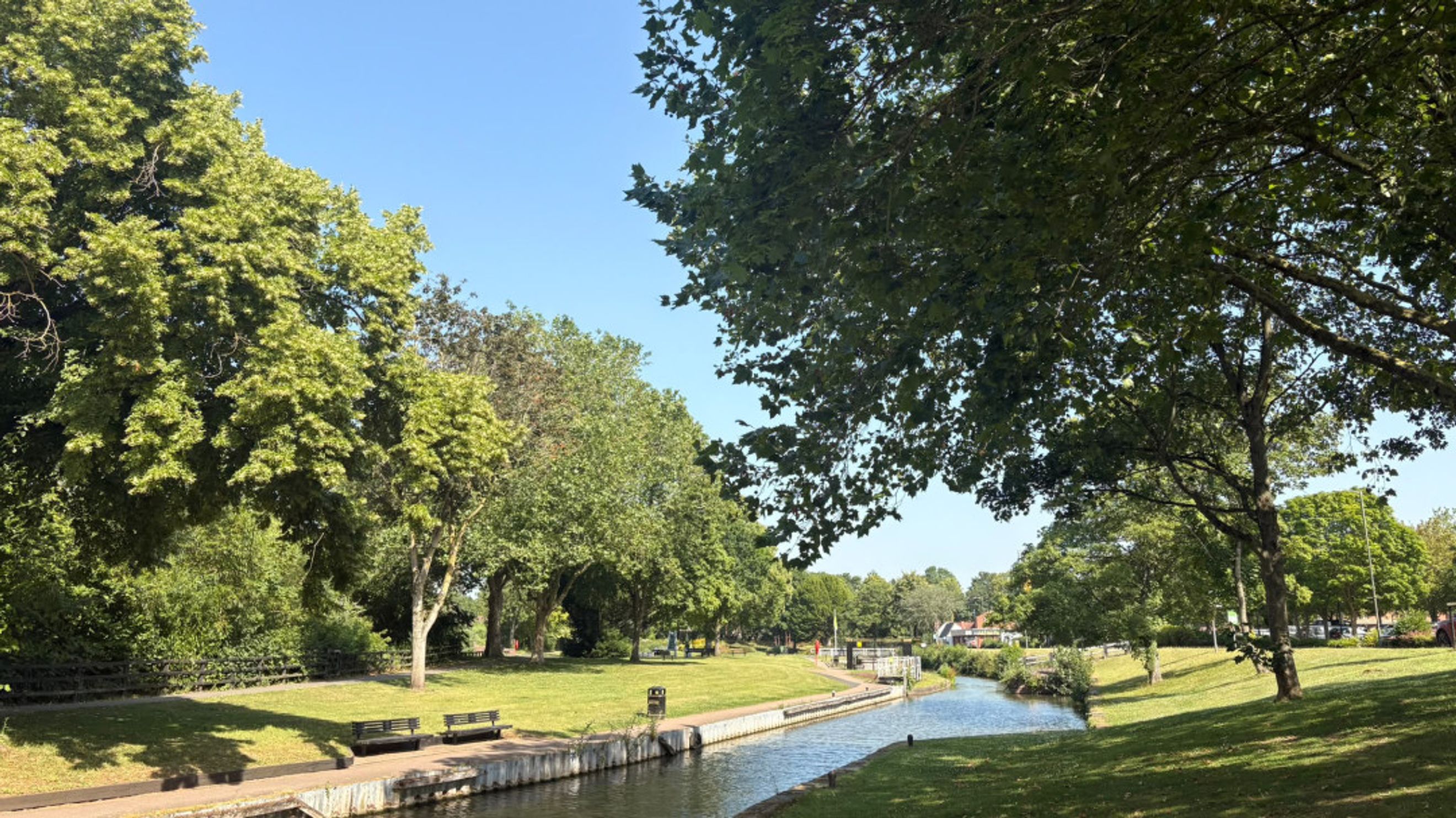 A canal runs through a park with trees and benches on a sunny day.