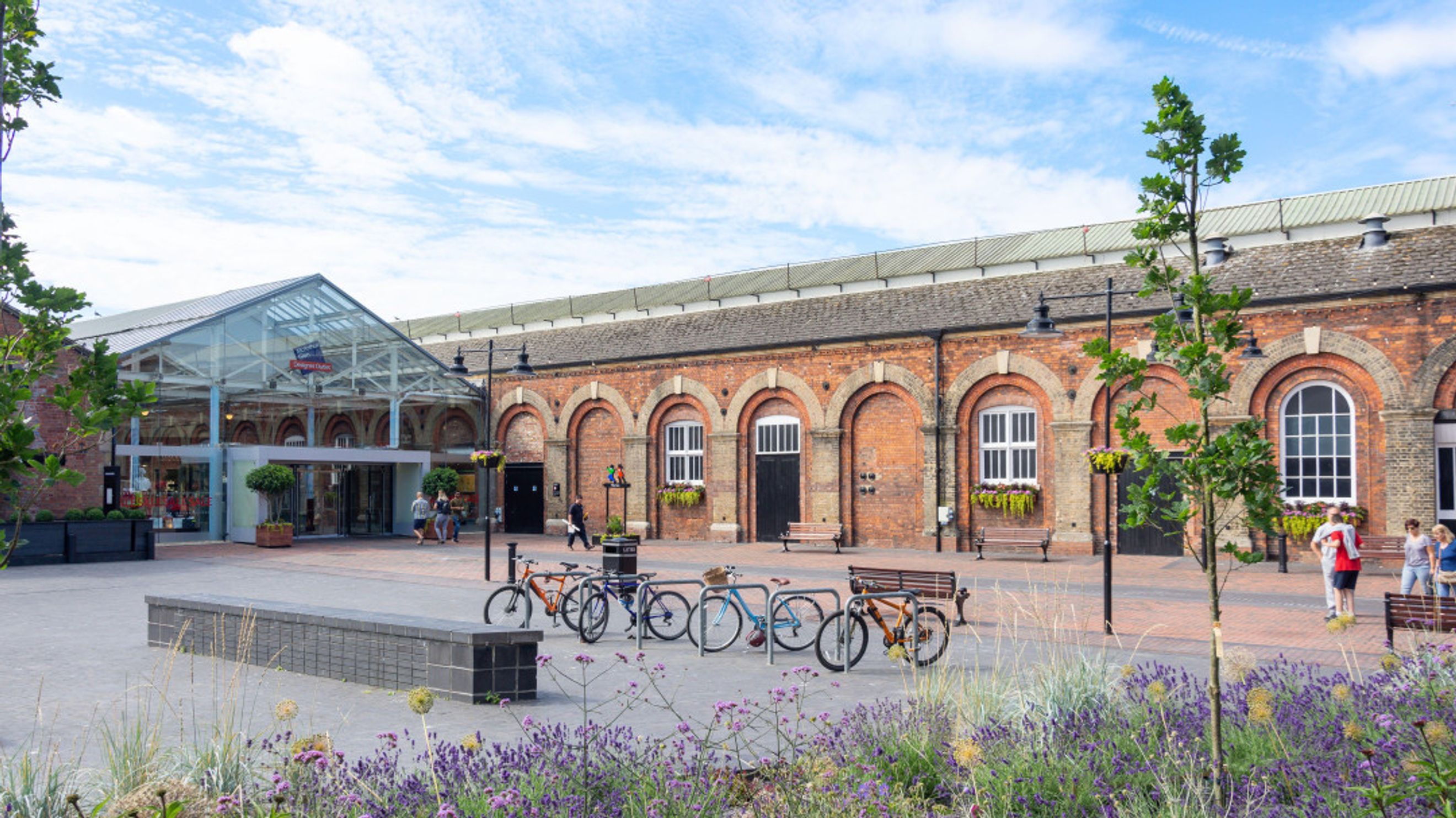 A courtyard with a glass-fronted building and brick architecture, featuring bicycle racks and people.