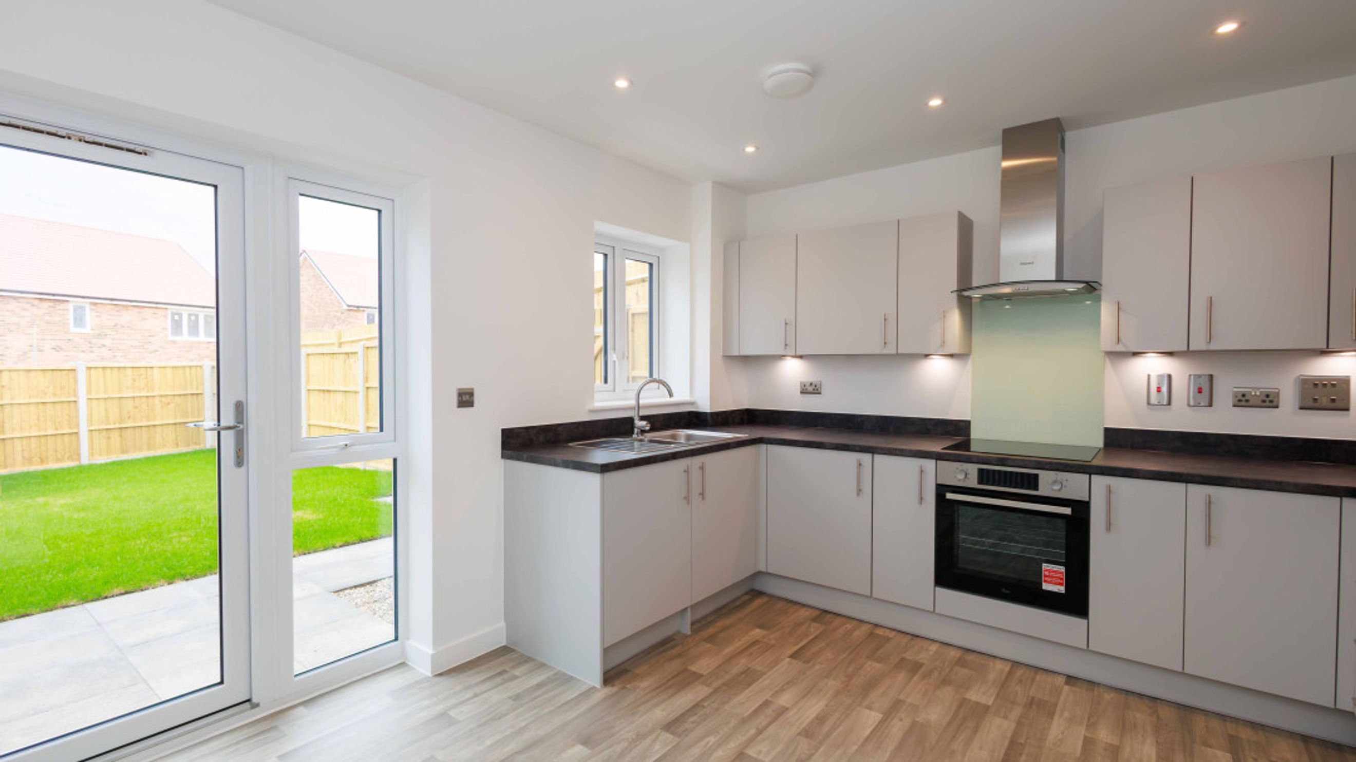 A modern kitchen with light gray cabinets, a dark countertop, a sink, and an oven, with double doors leading to a garden.