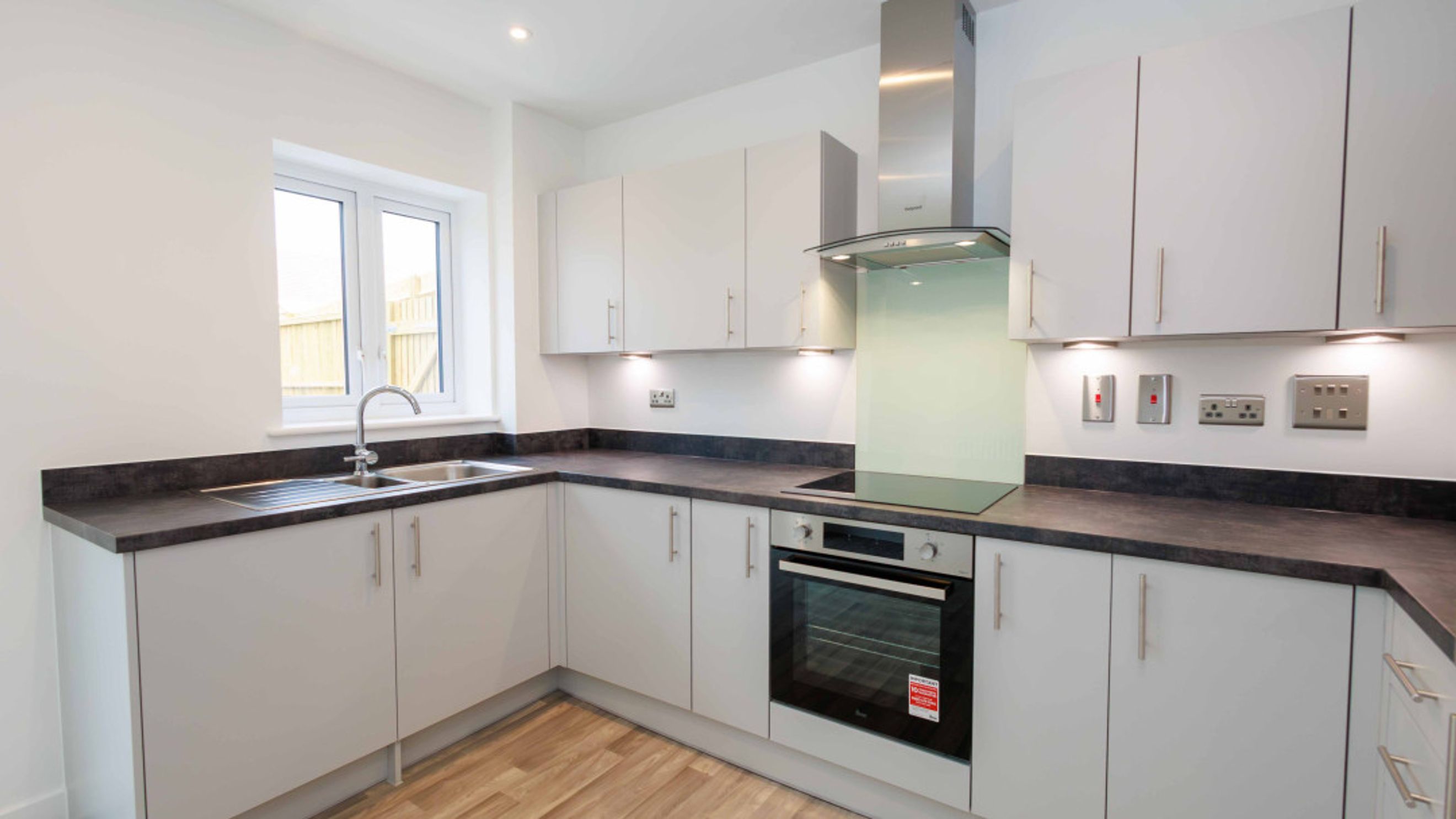 A modern kitchen with light gray cabinets, a dark countertop, a sink, an oven, and an extractor hood.
