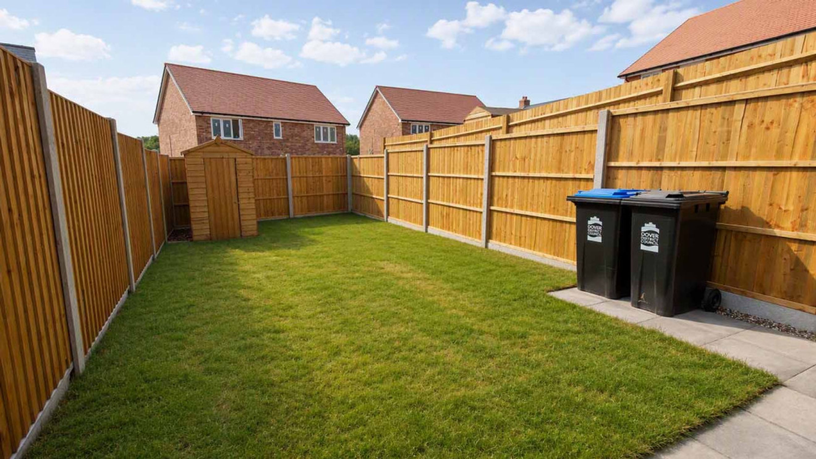 A backyard with a lawn, a wooden shed, a wooden fence, and two trash bins.