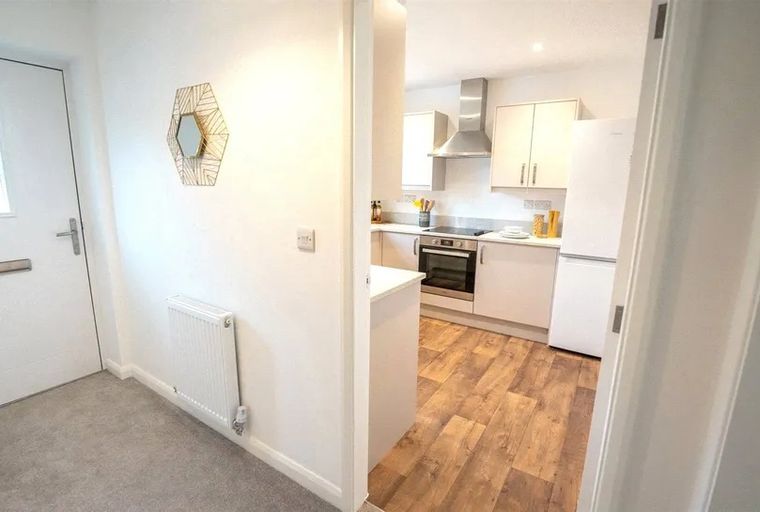 A hallway with a white door, a mirror, and a radiator, leading into a kitchen with white cabinets and a refrigerator.