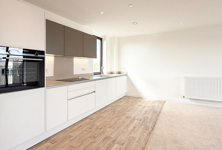 A modern kitchen with integrated appliances, handleless cabinets, and a stone countertop, adjacent to a carpeted living area with a radiator.