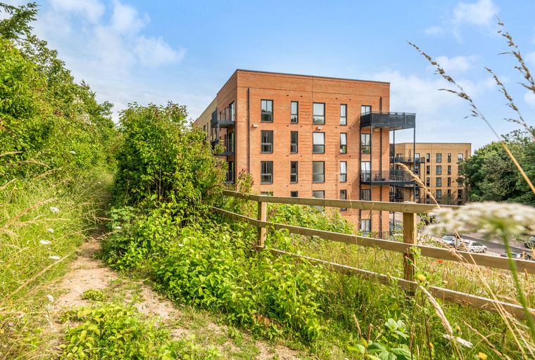 A modern brick apartment building with balconies, seen from a path through green foliage and a wooden fence.