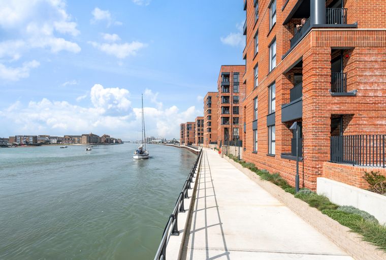A waterfront apartment building with a promenade and a river with a sailboat.