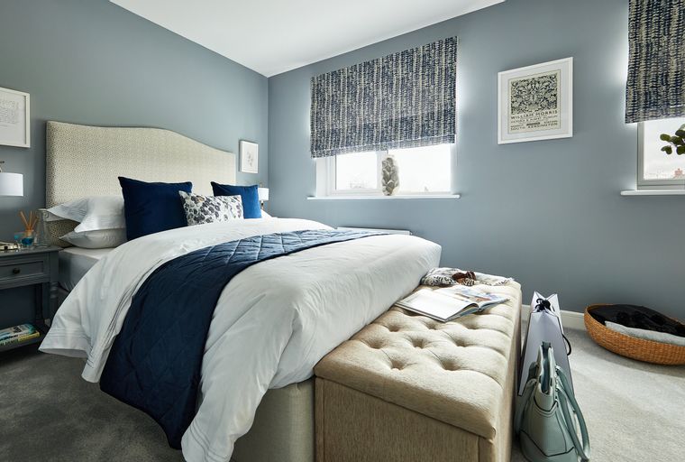 A bedroom with a bed, pillows, a headboard, a nightstand, a window with a blind, and an ottoman.