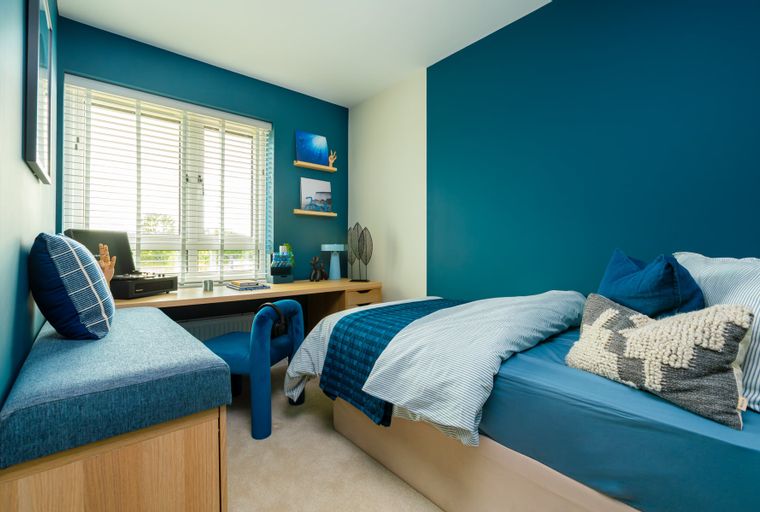 A bedroom with a bed, desk, chair, and window with blinds.