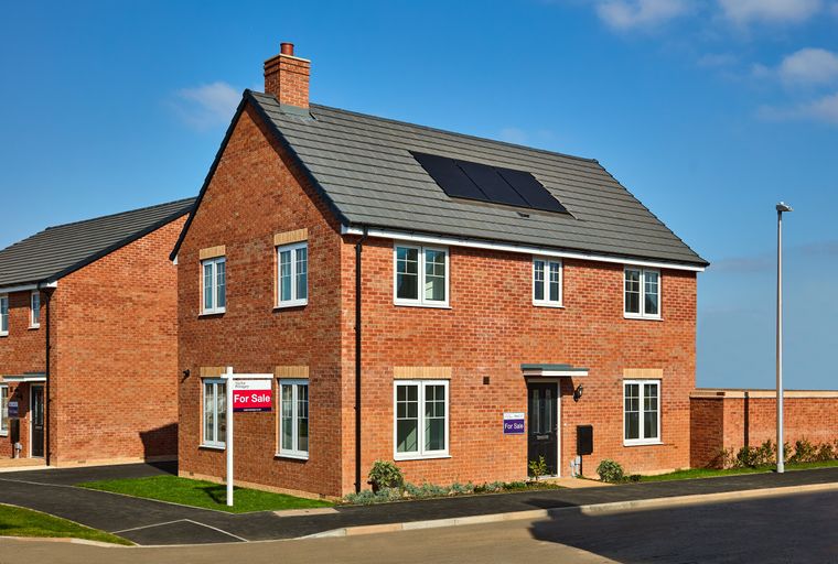 A red brick house with white-framed windows and solar panels on the roof, with a 'For Sale' sign in the front yard.