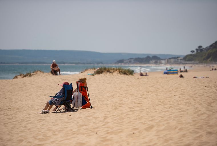A beach with two people sitting in chairs and a man sitting on a dune in the background.