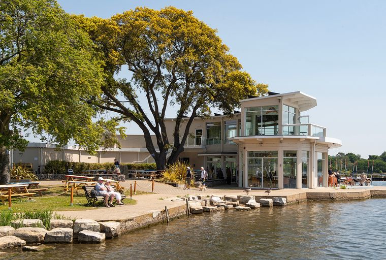 A modern building with glass windows situated by the water, with people and trees around.