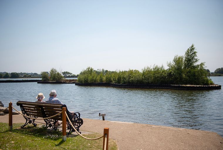 A couple sits on a bench overlooking a lake with islands of trees.