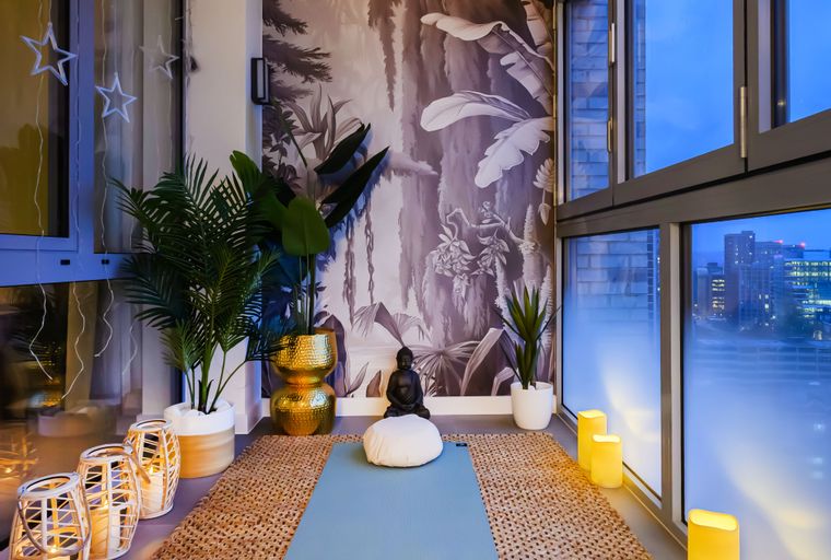 A yoga mat and meditation cushion are set up in a room with large windows, plants, and decorative lighting.