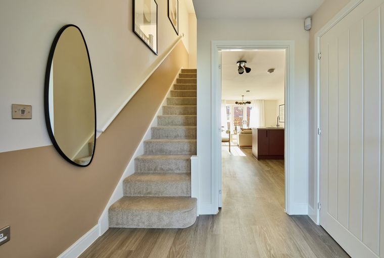 A hallway with a carpeted staircase, an oval mirror, and an open doorway leading to a living area.