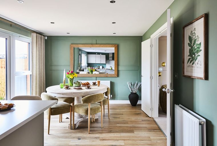 A dining area with a round table, chairs, and a mirror reflecting a kitchen.