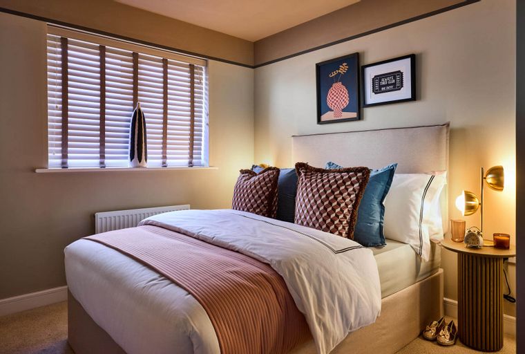A bedroom with a bed, pillows, a window with blinds, and a bedside table with a lamp.