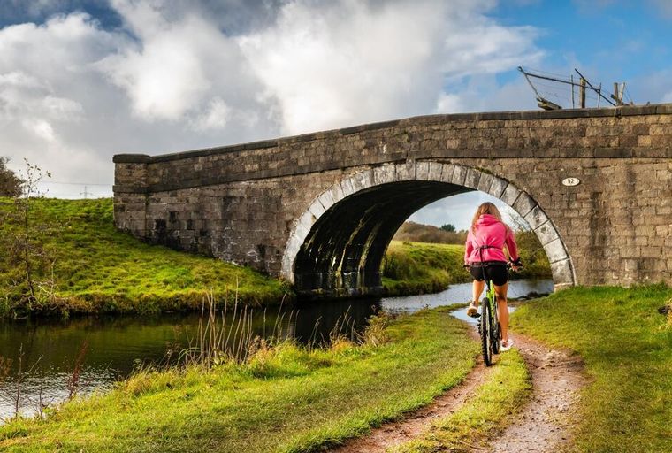 A person rides a bicycle on a dirt path under a stone bridge next to a canal.