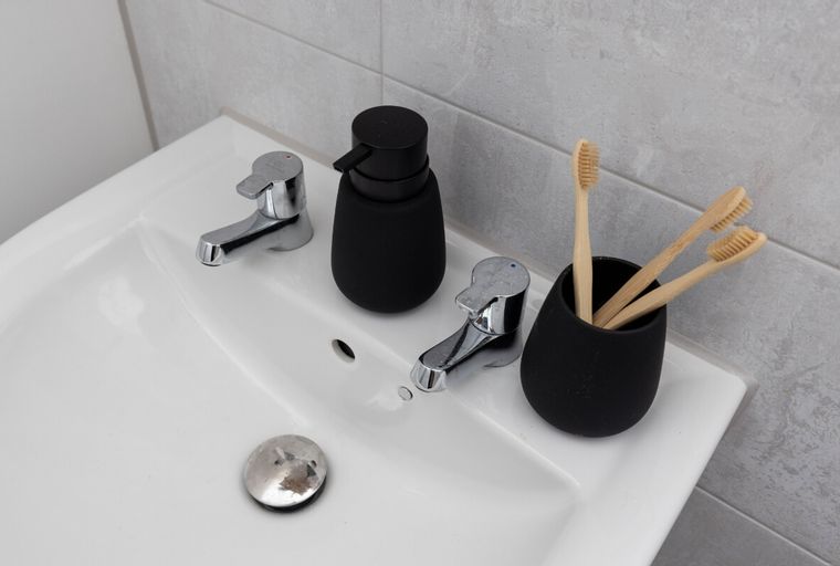 A white sink with two chrome faucets, a black soap dispenser, and a black cup holding three bamboo toothbrushes.