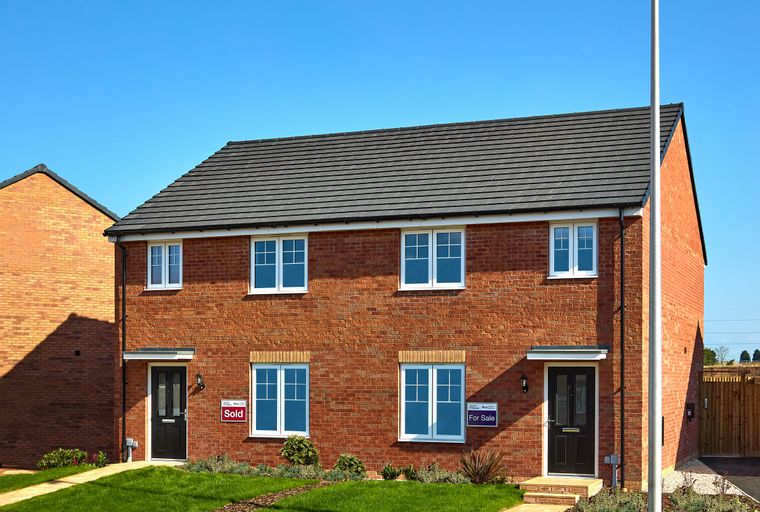 A pair of new red brick semi-detached houses with dark doors and white-framed windows under a grey tiled roof, set against a bright blue sky.