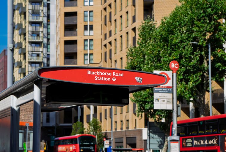 A bus stop sign for Blackhorse Road Station with a red double-decker bus in the background.