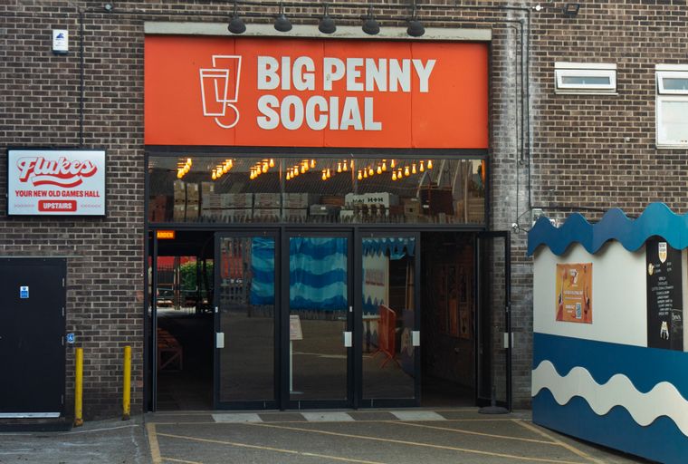 A brick building with an orange sign that reads 'BIG PENNY SOCIAL' and a 'Flukes' sign above a door.