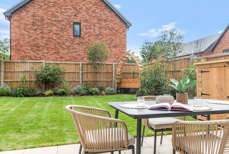 A patio table and chairs set up on a paved area in a garden with a lawn and a wooden fence.
