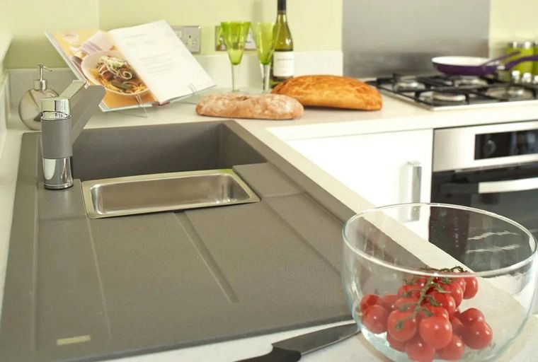 A kitchen counter with a sink, faucet, cookbook, bread, wine bottle, and tomatoes.
