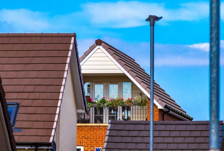A view of the upper floors of houses with brown tiled roofs and a balcony with flowers under a blue sky.