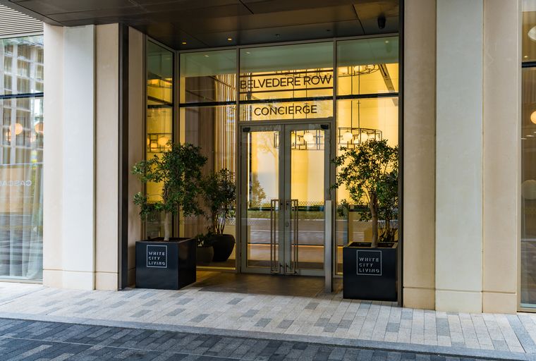 A modern building entrance with glass doors and signage for Belvedere Row Concierge, flanked by potted trees.